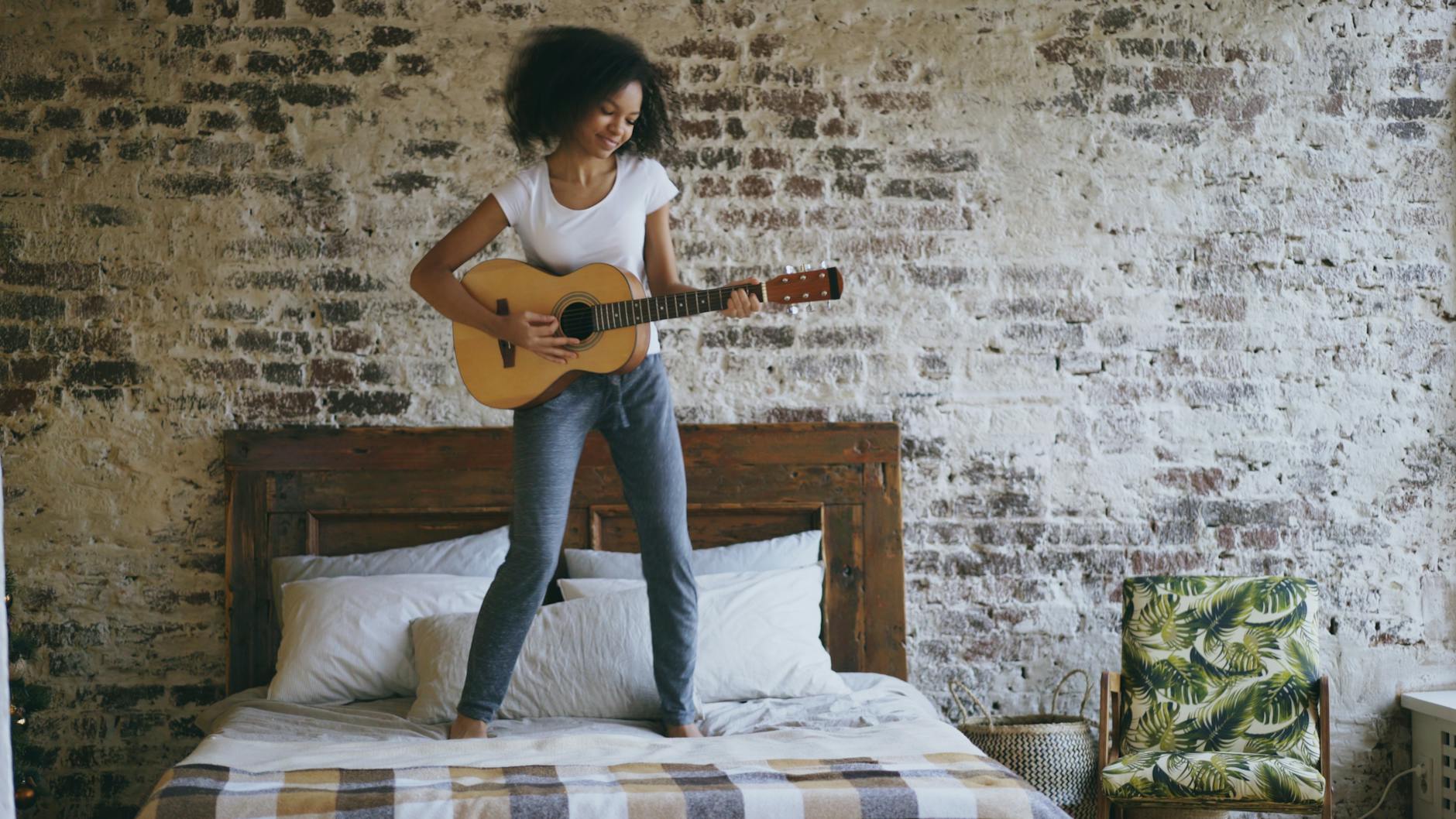 Młoda kobieta grająca na gitarze na łóżku w sypialni boho